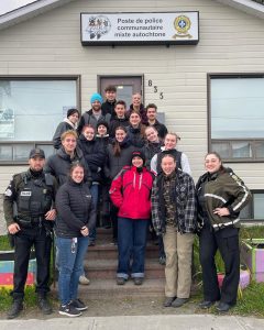 Groupe d'étudiantes et d'étudiants posant devant le poste de police communautaire mixte autochtone avec Nancy Moreau, enseignante et deux policiers.